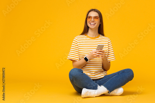 Happy young girl sitting on the floor, holding smartphone in hands and looking away, isolated on yellow background with copy space photo