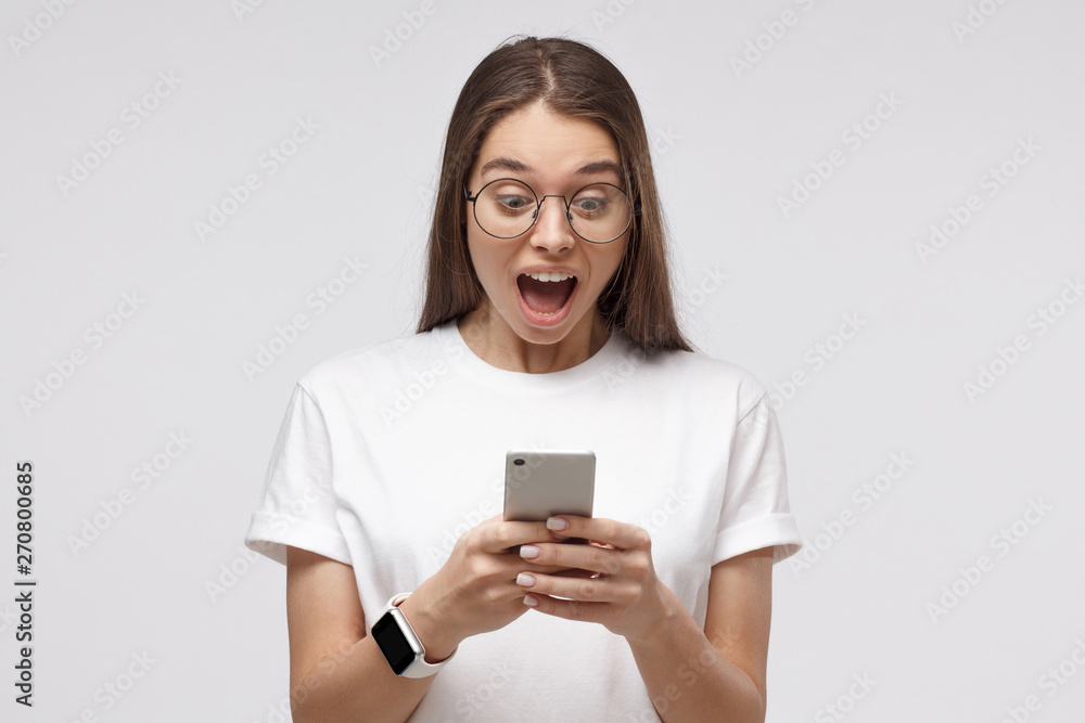 Close-up portrait of young female in white t-shirt, holding smartphone, looking surprised and shocked, isolated on gray background Close-up portrait of young female in white t-shirt, holding smartphone, looking surprised and shocked, isolated on gray background