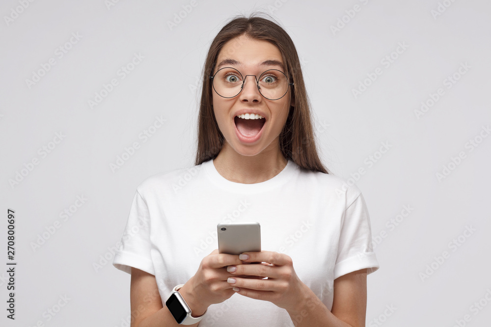 Wow! Close-up shot of young girl in white t-shirt, holding smartphone, looking surprised and shocked, isolated on gray background. Wow! Close-up shot of young girl in white t-shirt, holding smartphone, looking surprised and shocked, isolated on gray background.