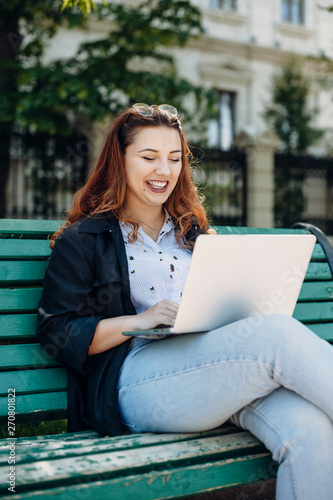 Portrait of a charming plus size woman sitting outside on a bench looking at her laptop on the legs smiling.