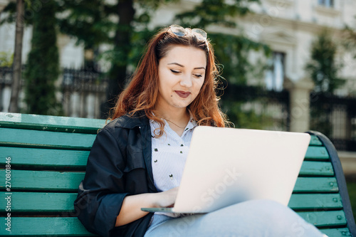 Young plus size caucasian female with red hair looking at her laptop smiling while sitting on a bench against a building.