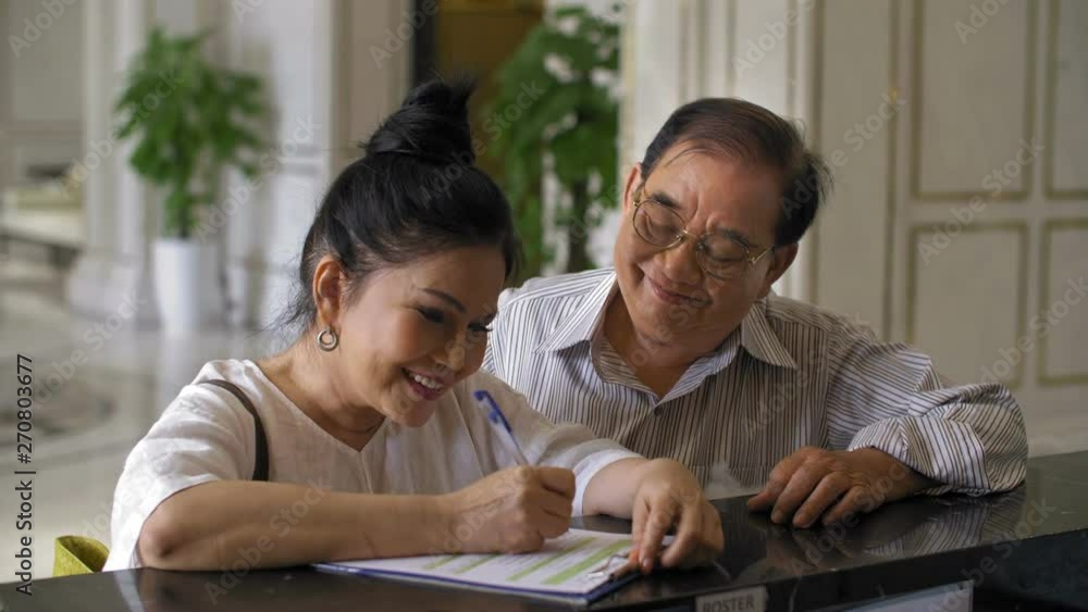 Lockdown of aged Asian woman filling in form at hotel reception and her spouse standing nearby Lockdown of aged Asian woman filling in form at hotel reception and her spouse standing nearby