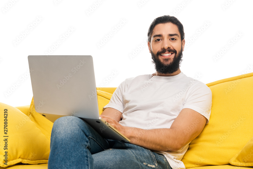 smiling latin man using laptop and sitting on sofa isolated on white smiling latin man using laptop and sitting on sofa isolated on white
