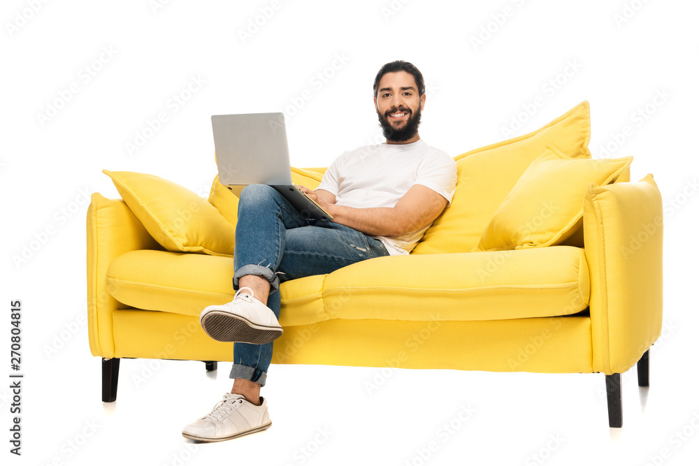 happy bearded latin man using laptop while sitting on yellow sofa isolated on white