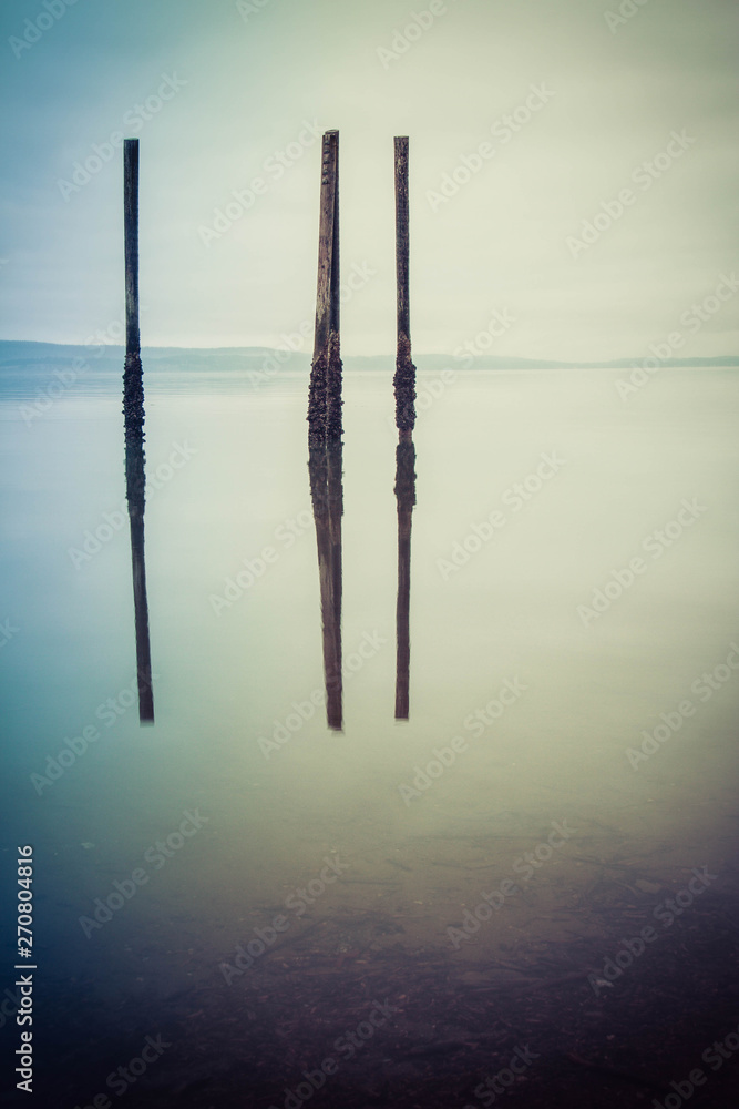 A gloomy image of the calm ocean with wood pilings standing free and reflecting in the water. A gloomy image of the calm ocean with wood pilings standing free and reflecting in the water.