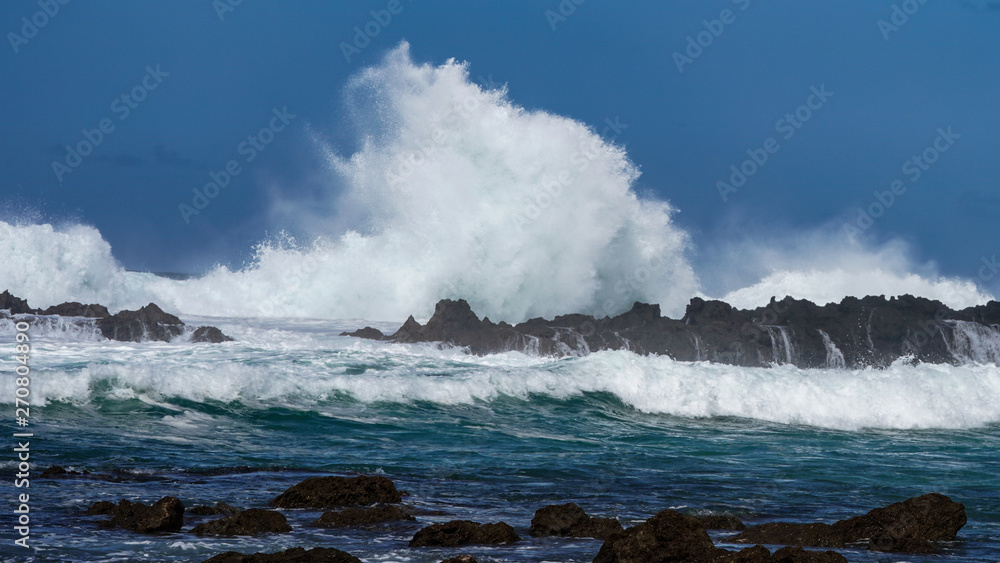 The angry sea, rough waters with a setting sun behind ominous clouds, emitting rays of light through the mist onto the angry sea, crashing wave upon the rocky shore.