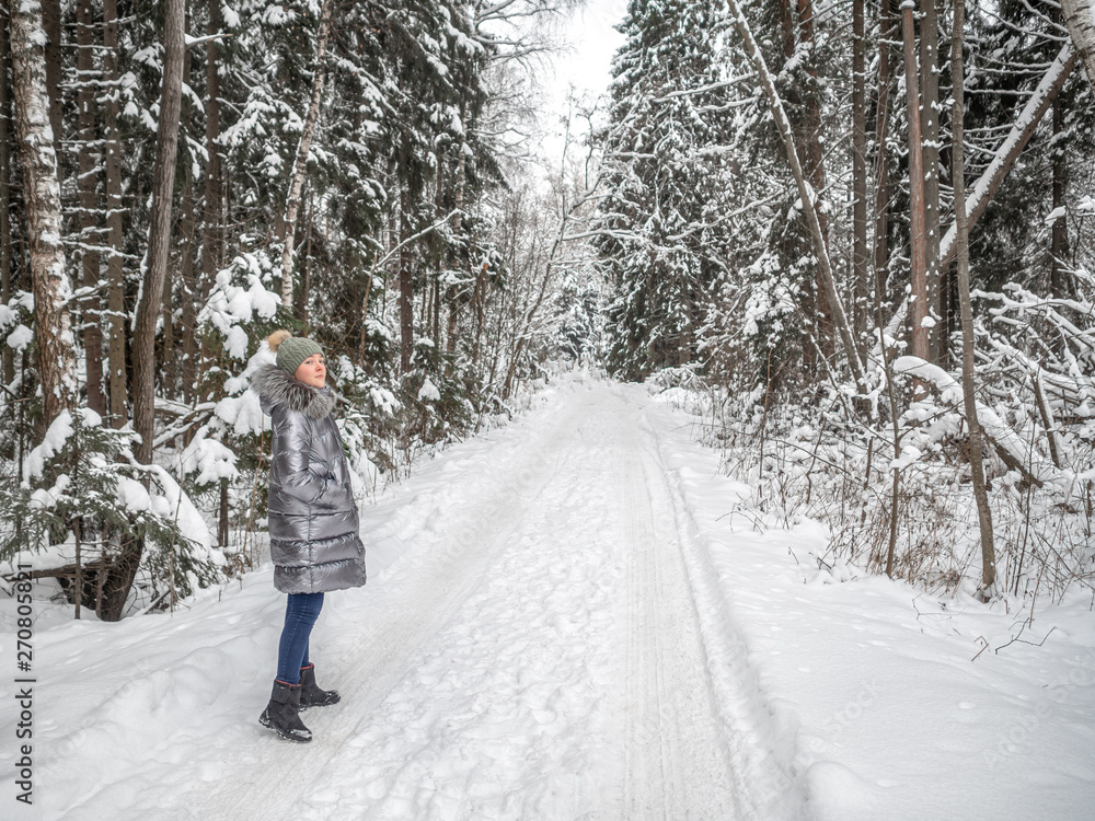 A young girl walks in the winter forest and enjoys silence. She is wearing a hat and a jacket of silver color