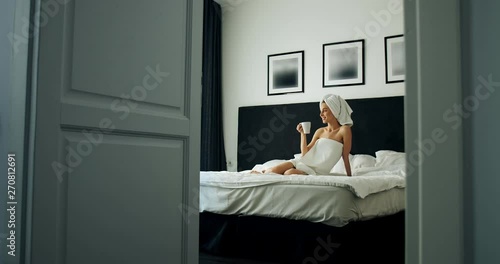 Wide shot of pretty woman dressed in towels drinking coffee sitting on the bed in stylish apartment bedroom