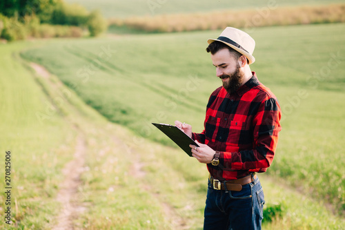 Young handsome bearded farmer with folder standing in green wheat field in early summer