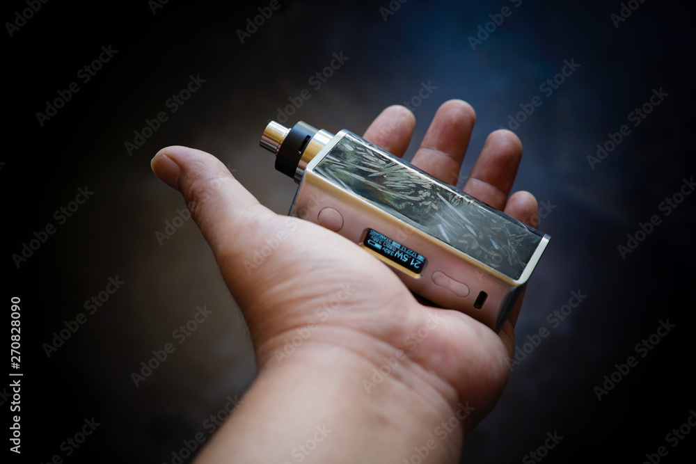 high end black carbon fiber in clear resin regulated box mods with rebuildable dripping atomizer and stainless drip tip in hand on dark grey texture background, vaporizer equipment, selective focus high end black carbon fiber in clear resin regulated box mods with rebuildable dripping atomizer and stainless drip tip in hand on dark grey texture background, vaporizer equipment, selective focus