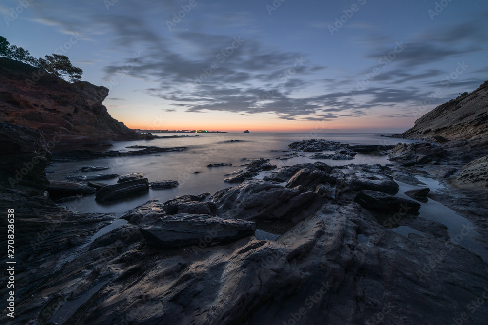 Rocks among the seashore at sunrise. There are cliffs at the sides under a cloudy sky.