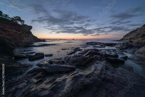 Rocks among the seashore at sunrise. There are cliffs at the sides under a cloudy sky. © Xavi
