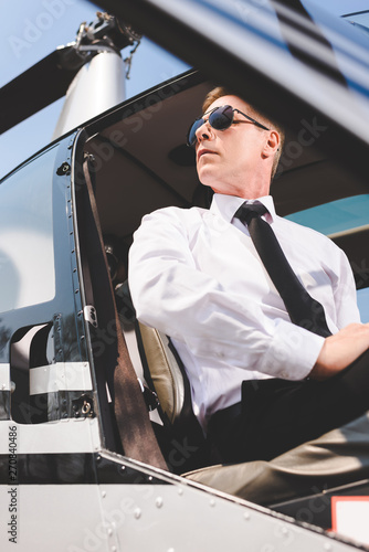 low angle view of handsome Pilot in sunglasses and formal wear sitting in helicopter cabin