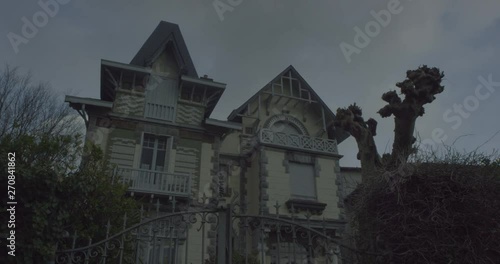 Establishing shot with tilt up of a haunted house in a creepy halloween atmosphere with an imposing old gate and a dead tree and green nature on the sides. Cloudy grey sky. Shot on RED EPIC DRAGON 6K.