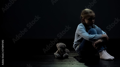 Little female kid sitting alone near teddy bear in dark room, lack of friends