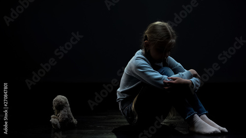 Sad little girl feeling lonely, sitting in dark room back to teddy bear toy