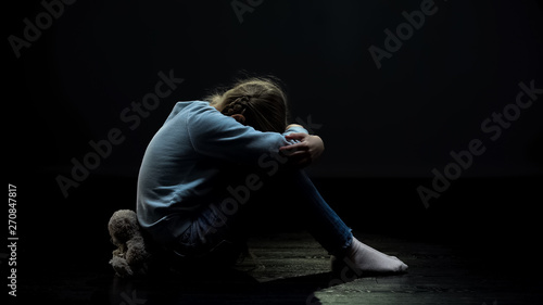 Upset little girl with teddy bear crying in dark abandoned room, loneliness