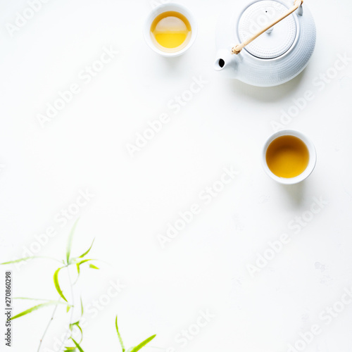 Asian tea concept, two white cups of tea and teapot surrounded with green tea dry leaves view from above, space for a text on white background.