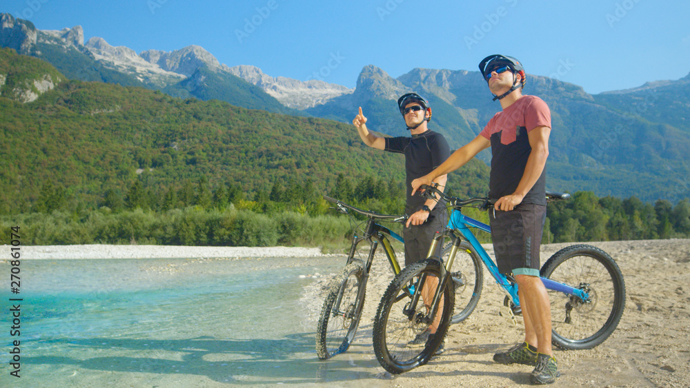 Fototapeta premium Young guys on ebikes stand by the river and look at the beautiful landscape.