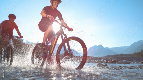 LOW ANGLE: Water drops fly around two guys riding electric bicycles along river. photo
