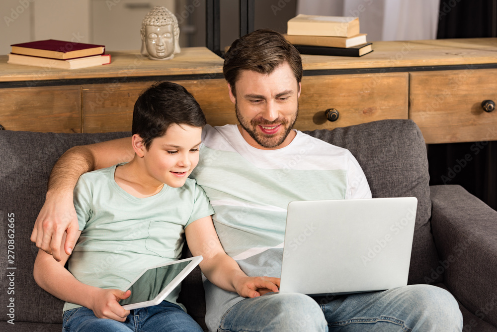son holding digital tablet and using laptop with father in living room son holding digital tablet and using laptop with father in living room