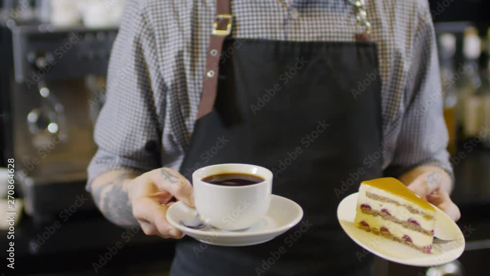Medium body shot of tattooed male barista walking up behind counter of coffee shop and standing in front of camera, holding Americano in white cup on saucer in one hand and slice of cake in another Medium body shot of tattooed male barista walking up behind counter of coffee shop and standing in front of camera, holding Americano in white cup on saucer in one hand and slice of cake in another