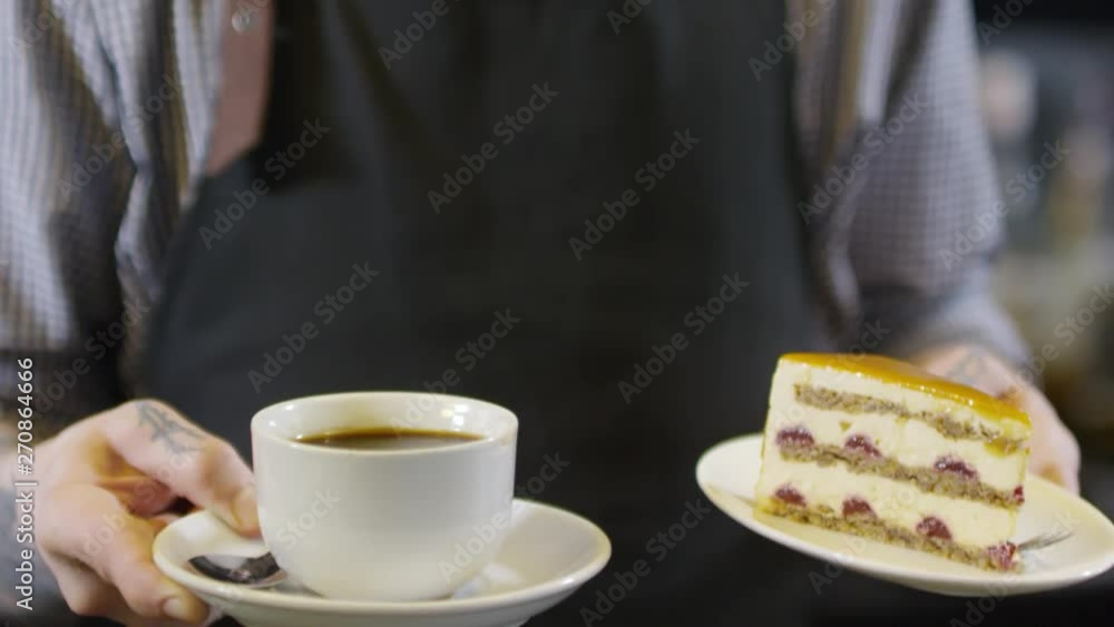 Close-up hands and object shot of tattooed male waiter standing in front of camera in café, lifting up and holding cup of Americano on white saucer and slice of cake on dessert plate Close-up hands and object shot of tattooed male waiter standing in front of camera in café, lifting up and holding cup of Americano on white saucer and slice of cake on dessert plate
