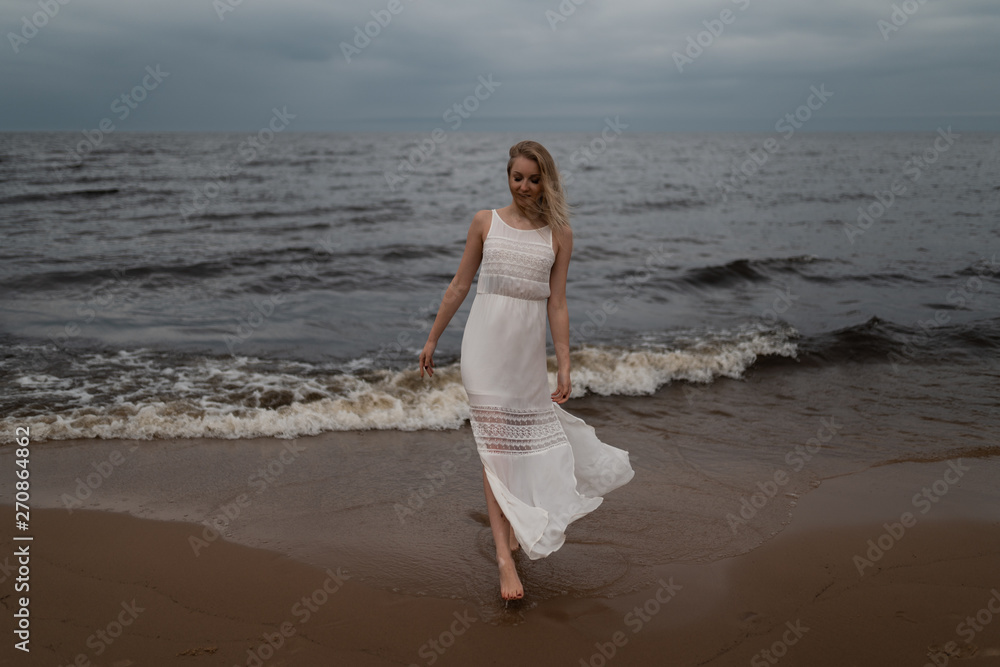 Walking Beautiful young blonde woman beach nymph in white dress near sea with waves during a dull gloomy weather with stormy wind and rain