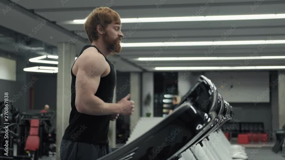 Redhead man running on treadmill machine in sport gym. Young bearded athletic man exercising and running on treadmill in sport gym with copy space Redhead man running on treadmill machine in sport gym. Young bearded athletic man exercising and running on treadmill in sport gym with copy space
