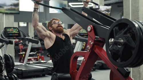 Redhead bodybuilder in glasses doing exercise on simulator in gym. Exercises for muscles of shoulders. Man with sports figure doing exercise on simulator in gym. Sport concept photo