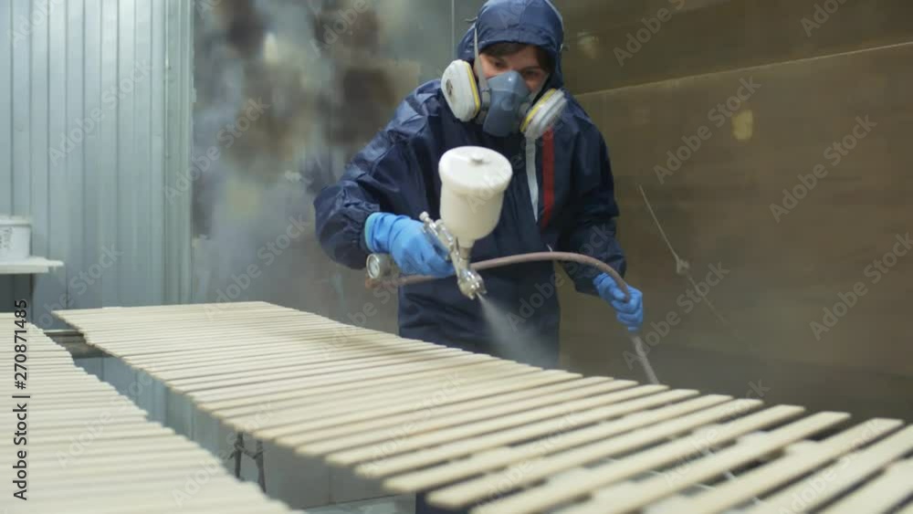 Man in respirator mask and protective costume painting wooden planks with spray gun in carpentry shop Man in respirator mask and protective costume painting wooden planks with spray gun in carpentry shop
