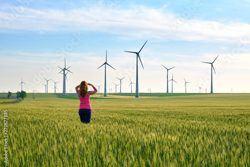 Obraz na płótnie Woman and wind turbines at sunset, in a field of green rye, with warm sun light. Concept for sustainable, renewable energy. w salonie