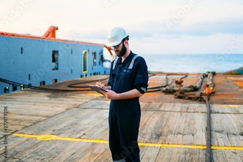 Deck Officer on deck of offshore vessel or ship , wearing PPE personal protective equipment. He fills checklist. Paperwork at sea photo