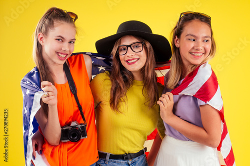 group of three laughing girls holding passports and tickets with photo camera wrapped in american flag spring party summer style yellow background studio, visa and independence day usa © yurakrasil