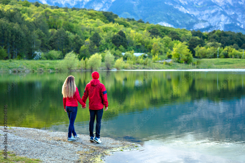 A couple in red jackets are standing and looking at the mountains with clouds, symmetrically reflected in the lake. Popular tourist attraction.