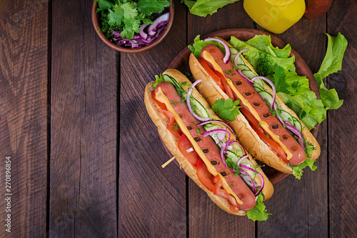 Hot dog with sausage, cucumber, tomato and lettuce on dark wooden background. Summer hotdog. Top view