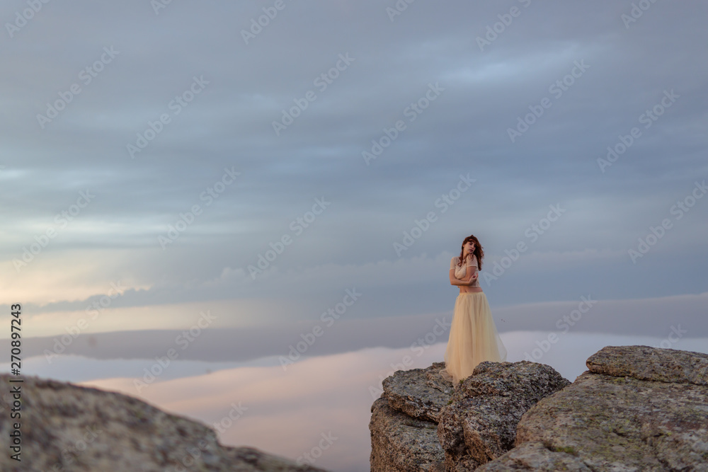A girl in a dress and hat admiring the dawn or sunset of the sun in a picturesque place in the mountains.