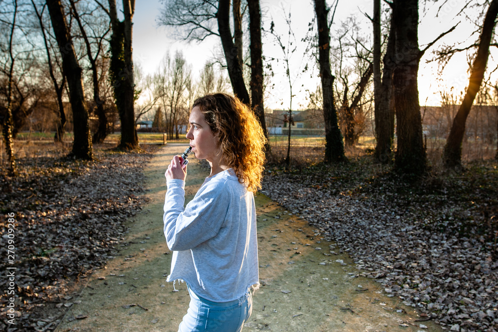 Young woman smoking electronic e-cigarette at the park on sunny day during sunset with smoke. Young woman smoking electronic e-cigarette at the park on sunny day during sunset with smoke.