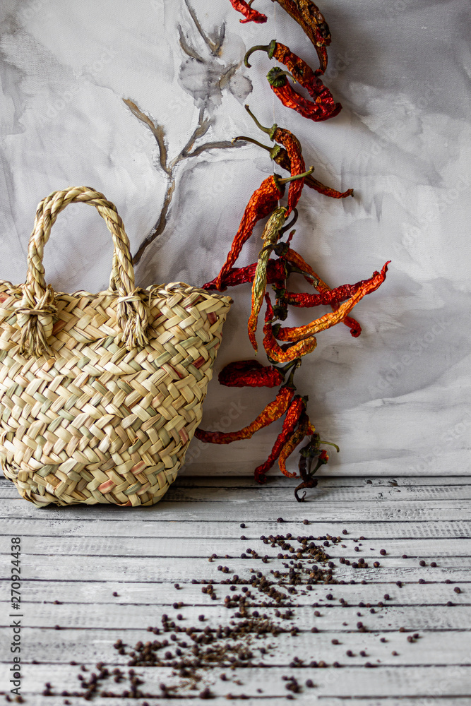 Dried red peppers on string with spreaded black pepper on wooden background with rush staw basket and cloth. Still life dried red and black pepper at old kitchen with handbag.