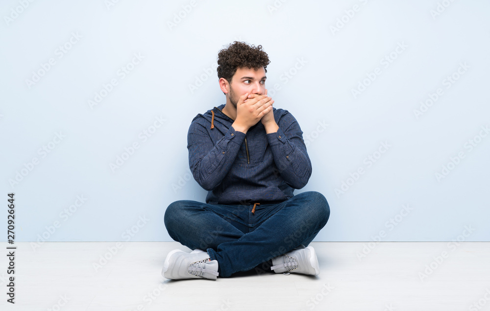 Young man sitting on the floor covering mouth and looking to the side Young man sitting on the floor covering mouth and looking to the side