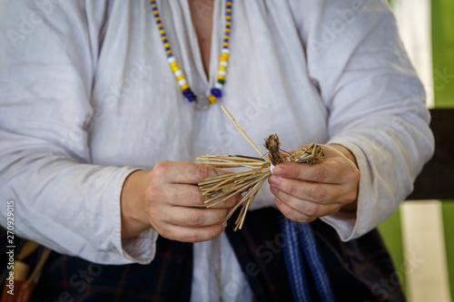 Woman in Russian ethnic clothes makes a small toy of straw in shape of man