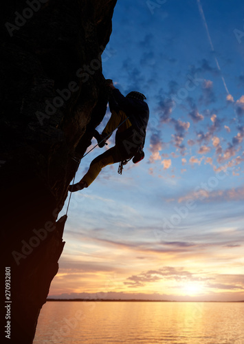 Silhouette of sport man climbing up to top of mountain summit. Sunset with blue cloudy sky over river and copy space on background. Healthy lifestyle, goal achievement and adventure active concept.