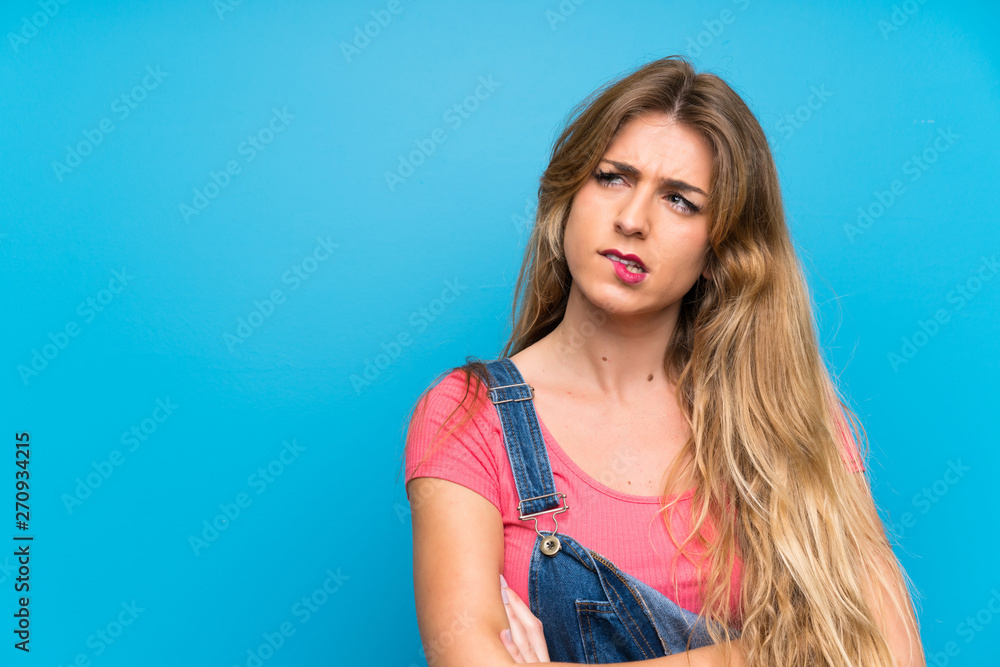 Young blonde woman with overalls over isolated blue wall with confuse face expression Young blonde woman with overalls over isolated blue wall with confuse face expression