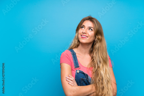 Young blonde woman with overalls over isolated blue wall looking up while smiling