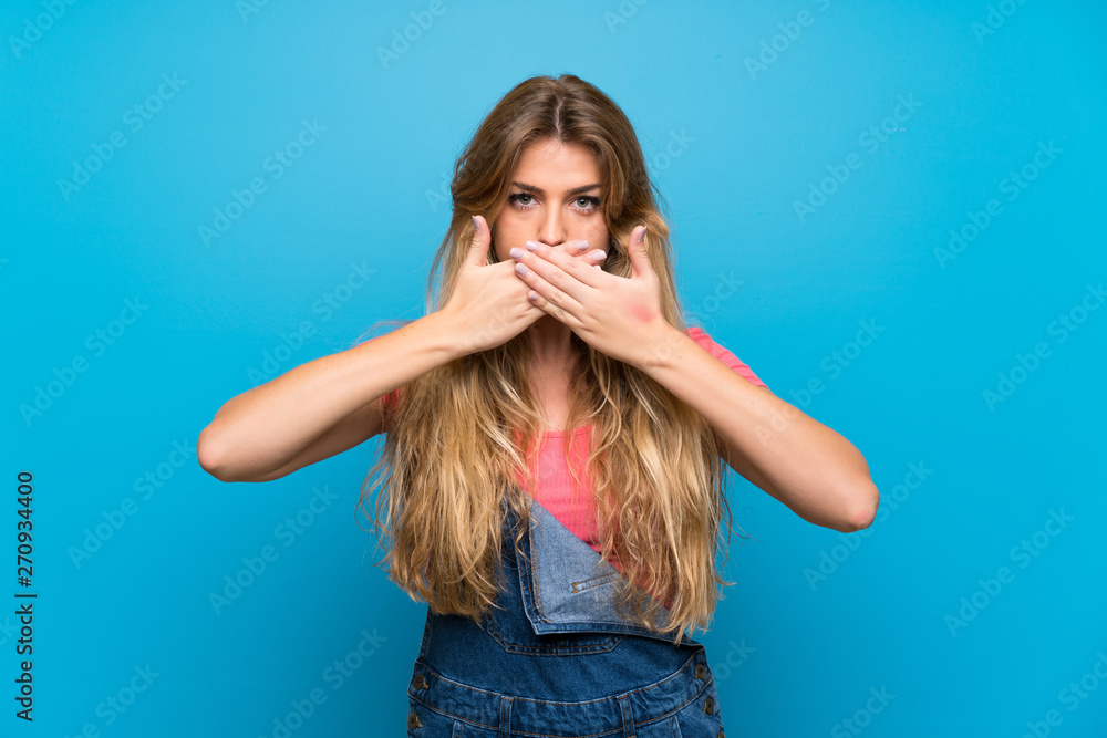 Young blonde woman with overalls over isolated blue wall covering mouth with hands Young blonde woman with overalls over isolated blue wall covering mouth with hands