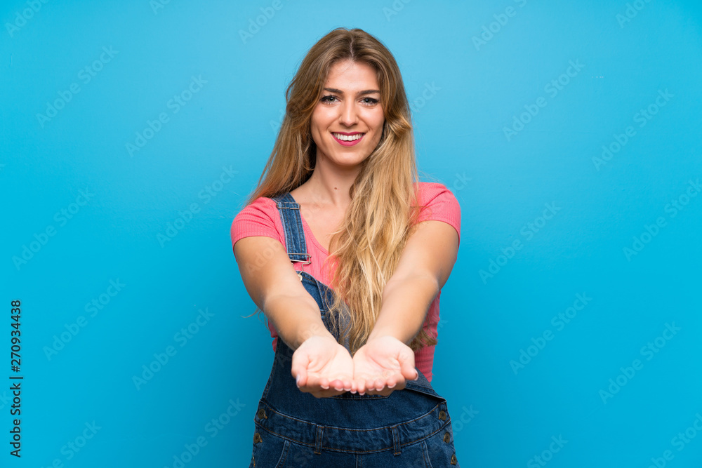 Young blonde woman with overalls over isolated blue wall holding copyspace imaginary on the palm to insert an ad Young blonde woman with overalls over isolated blue wall holding copyspace imaginary on the palm to insert an ad
