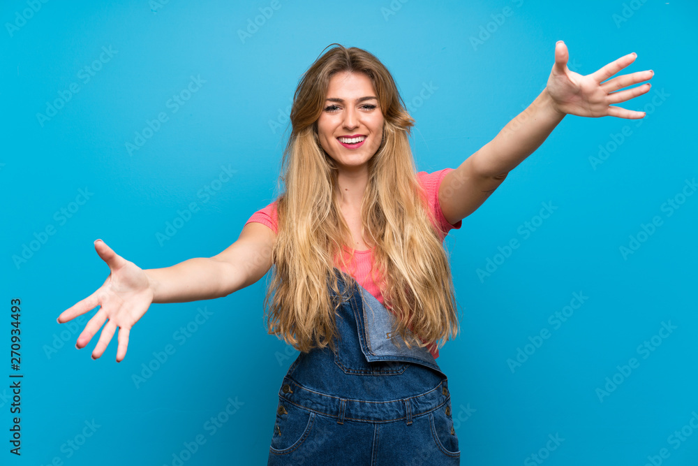 Young blonde woman with overalls over isolated blue wall presenting and inviting to come with hand Young blonde woman with overalls over isolated blue wall presenting and inviting to come with hand