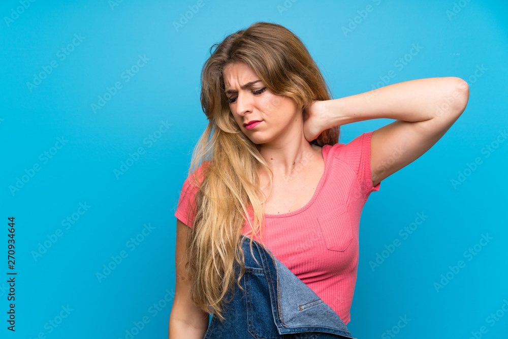 Young blonde woman with overalls over isolated blue wall with neckache Young blonde woman with overalls over isolated blue wall with neckache
