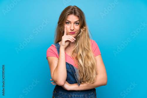 Young blonde woman with overalls over isolated blue wall Looking front