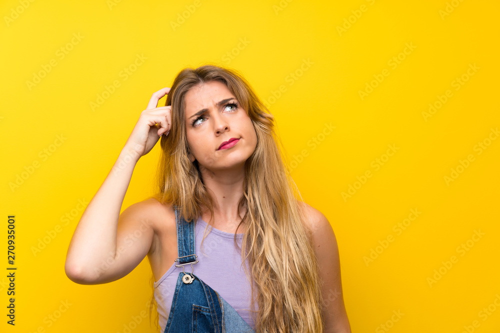 Young blonde woman with overalls over isolated yellow background having doubts and with confuse face expression
